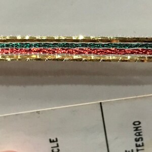May include: Close-up of decorative ribbon featuring gold, green, and red strands. The ribbon has a metallic sheen and is likely used for crafts or holiday decorations. The ribbon is layered with a gold border.