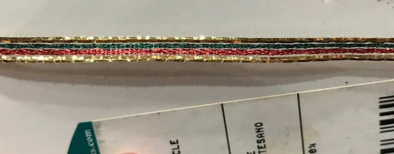 May include: Close-up of decorative ribbon featuring gold, green, and red strands. The ribbon has a metallic sheen and is likely used for crafts or holiday decorations. The ribbon is layered with a gold border.