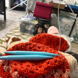 May include: A close-up shot of knitting equipment. Two light blue knitting needles rest on a bright orange and cream crocheted item. A ball of yarn and a clear plastic cable are also present, indicating a crafting project.