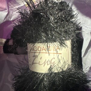 May include: A spool of black novelty yarn with metallic strands. The label reads "MODA DE LA ZING". The yarn is fluffy and has a textured appearance, suitable for various craft projects.