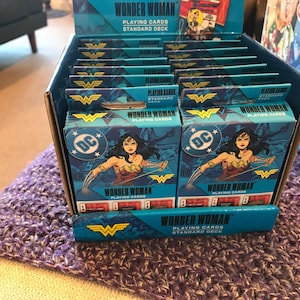 May include: A display box filled with Wonder Woman playing card decks. Each deck features the DC logo and an image of Wonder Woman. The box and decks have a teal and gold color scheme, with the words "Wonder Woman" and "Playing Cards" visible.