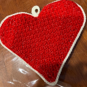 May include: A handmade, red crocheted heart-shaped potholder with a white border and a loop for hanging. The potholder is made of a textured yarn and is suitable for kitchen use.
