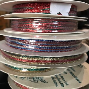 May include: A stack of spools of decorative ribbon in various colors. The ribbons include red, green, blue, silver, and gold metallic threads. The spools are white with text in French and Spanish.