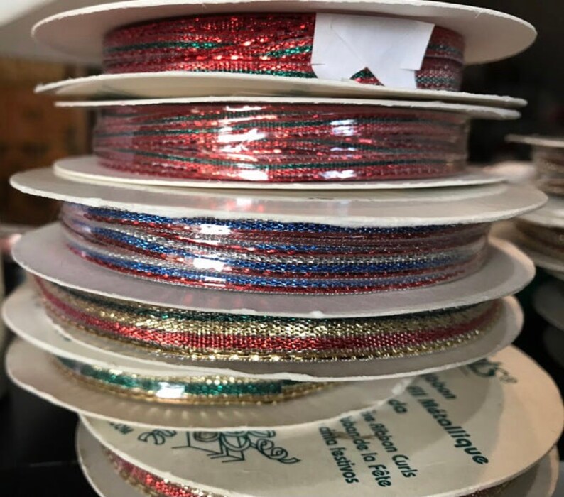 May include: A stack of spools of decorative ribbon in various colors. The ribbons include red, green, blue, silver, and gold metallic threads. The spools are white with text in French and Spanish.