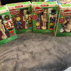 May include: Four boxed wood paint sets featuring Christmas-themed ornaments. The sets include a Nutcracker Drummer, Nutcracker Guard, and Snowman. Each set contains a wooden ornament and four acrylic paints, perfect for holiday crafting.