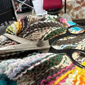 Puede incluir: Unas tijeras con mango negro, marcadas "Sta-Sharp Made in USA", descansan sobre un textil colorido tejido a mano. El textil presenta una variedad de colores, incluyendo marrón, blanco, amarillo, rosa y azul, creando un patrón texturizado y multicolor.