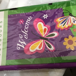 Könnte beinhalten: Eine violette Willkommens-Gartenflagge mit grünem Rand. Die Flagge zeigt zwei bunte Schmetterlinge mit gelben, roten, orangefarbenen, rosa und grünen Akzenten. Der Text "Willkommen" ist in einer dekorativen Schrift geschrieben.