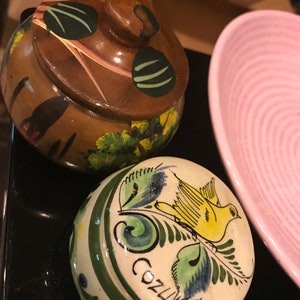 May include: Two small, round, ceramic trinket boxes with painted designs. One box is brown with green and black accents and a lid. The other box is white with green, blue, and yellow accents and a bird design. The word "Cozumel" is written on the box.