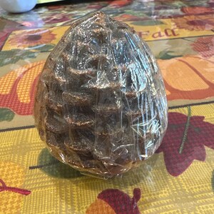 May include: A large, brown, pinecone-shaped decorative item wrapped in clear plastic. The pinecone has a textured surface and is sitting on a fall-themed tablecloth with autumn leaves and acorn designs.