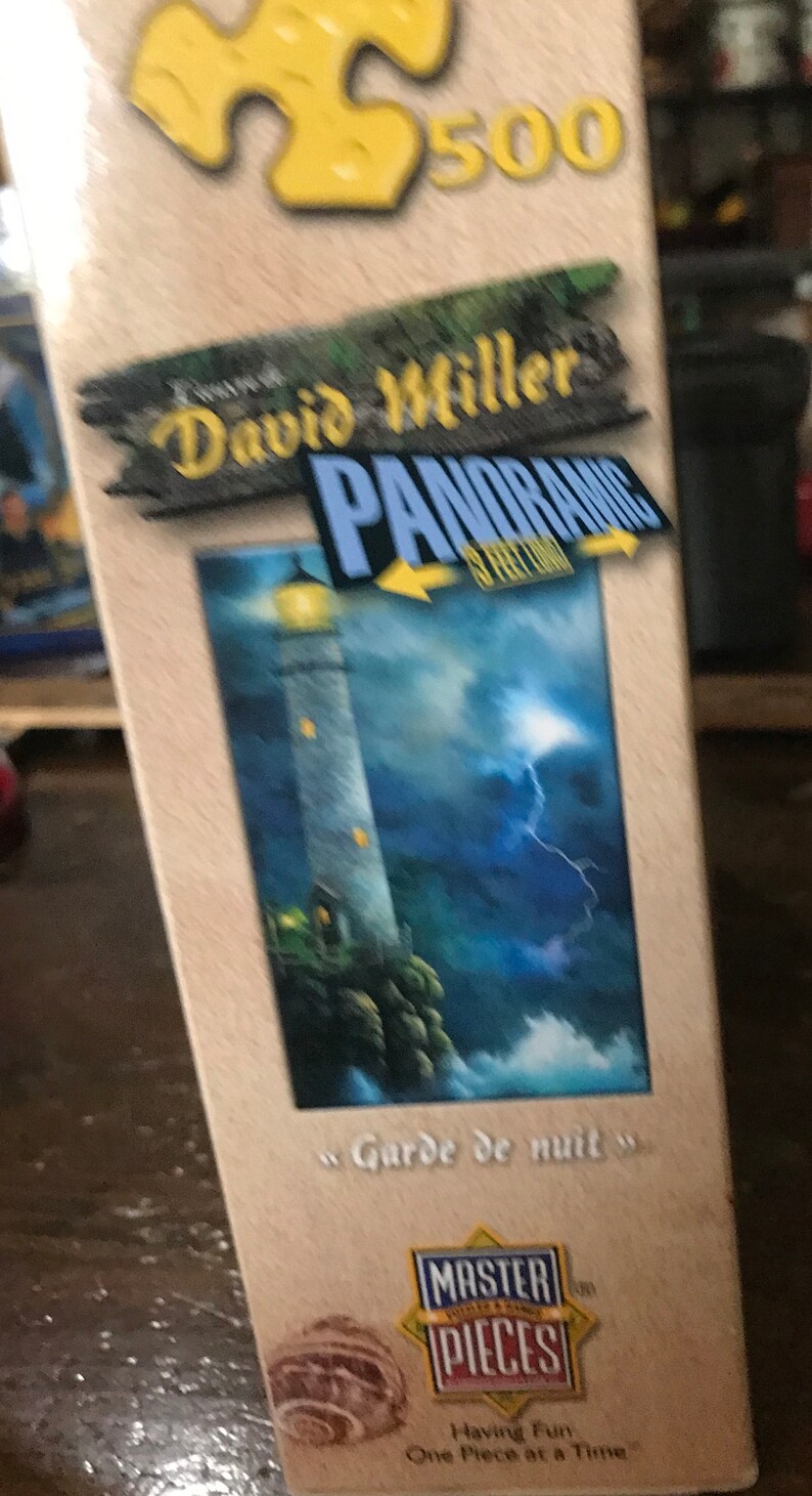 May include: A 500-piece jigsaw puzzle box featuring a painting of a lighthouse with a lightning bolt in the sky. The box is beige with a yellow puzzle piece in the top left corner. The title of the puzzle is "David Miller Panoramic" and the painting is titled "Garde de nuit". The box also features the Master Pieces logo.