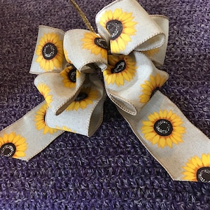 May include: A decorative bow with a sunflower pattern on a light grey ribbon. The sunflowers have yellow petals and dark brown centres. A gold-coloured wire stem is attached to the bow. The bow is resting on a purple textured surface.