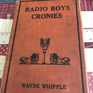 May include: A vintage book titled "Radio Boys Cronies" by Wayne Whipple. The cover is a reddish-brown color with black text and illustrations, including two lions and an emblem. The book is rectangular and appears to be well-worn.