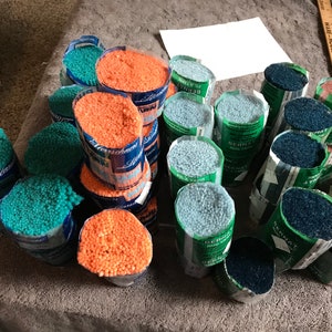 May include: A variety of yarn spools in different colors, including teal, orange, blue, and brown. The spools are arranged in a pile on a gray surface. The yarn is labeled "Bernat Rug Yarn" and "Sternschnuppchen"