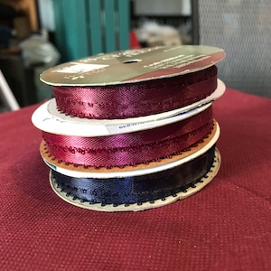 May include: A stack of spools of satin ribbon in shades of burgundy and navy blue. Each spool has a decorative edge and is made of a smooth, shiny material. The ribbon is likely used for crafts, gift wrapping, or other decorative purposes.