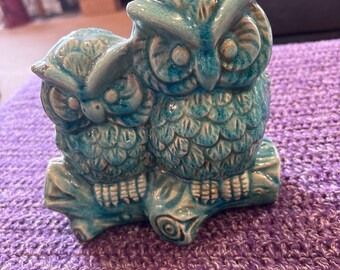 May include: A ceramic owl figurine in turquoise glaze. The figurine depicts two owls perched together on a branch. The owls have detailed feather patterns and large eyes. The figurine is placed on a purple knitted surface.