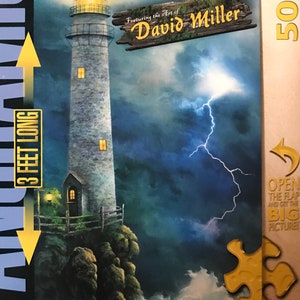 May include: A 500-piece jigsaw puzzle featuring a lighthouse with a light on top, a door, and windows. The lighthouse is on a rocky cliff with a wooden fence. The sky is dark blue with a lightning bolt and clouds. The puzzle box has the text "Featuring the Art of David Miller" and "3 feet long".
