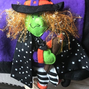 May include: A green witch doll with orange hair, a black hat, and a black and white striped dress holding a brown broom. The doll is on a purple and black background with white stars.