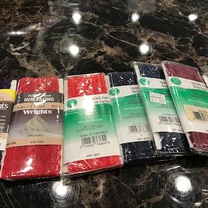 May include: A close-up of eight packages of stretch lace hem facing in various colors, including red, navy, and burgundy. The packages are labeled with the brand name "Wrights" and the product name "Stretch Lace Hem Facing".
