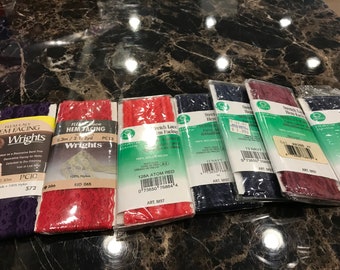 May include: A close-up of eight packages of stretch lace hem facing in various colors, including red, navy, and burgundy. The packages are labeled with the brand name "Wrights" and the product name "Stretch Lace Hem Facing".