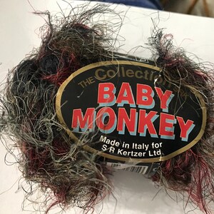 May include: A skein of yarn in a mix of black, grey, and red colours. The yarn is very fluffy and has a fuzzy texture. The label reads "The Collection Baby Monkey Made in Italy for S.R. Kertzer Ltd."