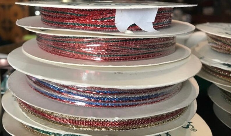 May include: Spools of decorative ribbon in various colors, including red, blue, and gold. The ribbon is metallic and has a woven texture. The spools are stacked on top of each other, creating a visually appealing display. Perfect for crafting and gift wrapping.