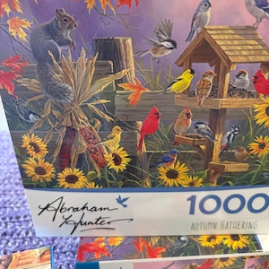 May include: A 1000-piece jigsaw puzzle featuring a detailed illustration of an autumn scene. The artwork depicts a variety of colorful birds gathered around a bird feeder, with a squirrel, sunflowers, and fall foliage. The puzzle is labeled "Autumn Gathering."