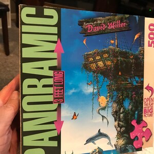 May include: A 500-piece panoramic jigsaw puzzle featuring the art of David Miller. The puzzle depicts a whimsical scene with a wooden structure, a mermaid, dolphins, and butterflies against a blue sky. The puzzle box is 3 feet long.