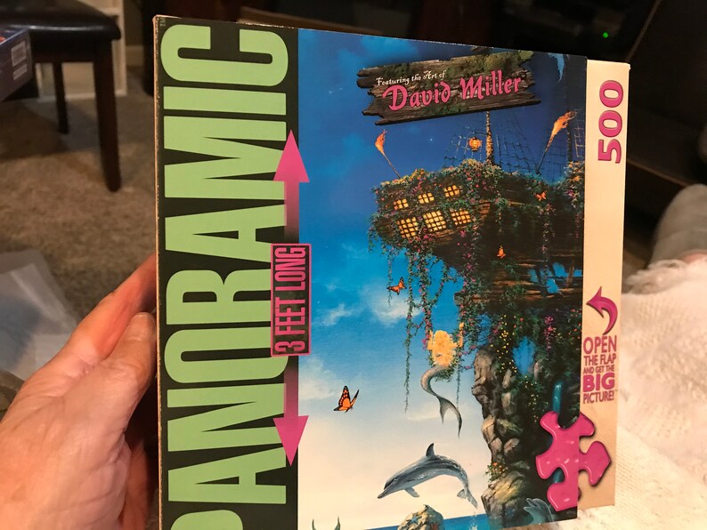 May include: A 500-piece panoramic jigsaw puzzle featuring the art of David Miller. The puzzle depicts a whimsical scene with a wooden structure, a mermaid, dolphins, and butterflies against a blue sky. The puzzle box is 3 feet long.