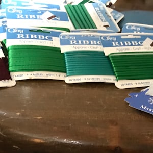 Könnte beinhalten: Sortiment an grünen, blaugrünen und burgunderfarbenen Bandspulen, jede mit der Aufschrift "Offray Ribbon" und der Länge und Breite des Bandes. Ein Paket mit weißer Spitzenborte ist ebenfalls enthalten, beschriftet mit "Winter Classic Snowflake Trim" und "Makes 3 Snowflakes".