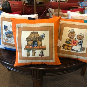 May include: Five decorative throw pillows with a Thanksgiving theme. The pillows feature a variety of designs, including a birdhouse with pumpkins, a pilgrim, and two bears holding a heart.