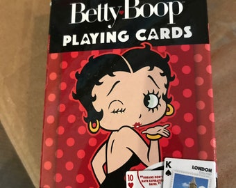 Peut inclure: Un jeu de cartes à jouer Betty Boop. La boîte est noire avec les mots "Betty Boop PLAYING CARDS" en blanc. Le fond est rouge avec des pois blancs. La boîte présente une illustration de Betty Boop faisant un bisou. Les cartes présentent des motifs personnalisés.