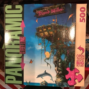 May include: A 500-piece panoramic jigsaw puzzle featuring the art of David Miller. The puzzle depicts a whimsical scene with a mermaid, a dolphin, butterflies, and a treehouse-like structure on a cliff overlooking the ocean. The box is green and black with the words "Panoramic" and "3 feet long" printed on it.