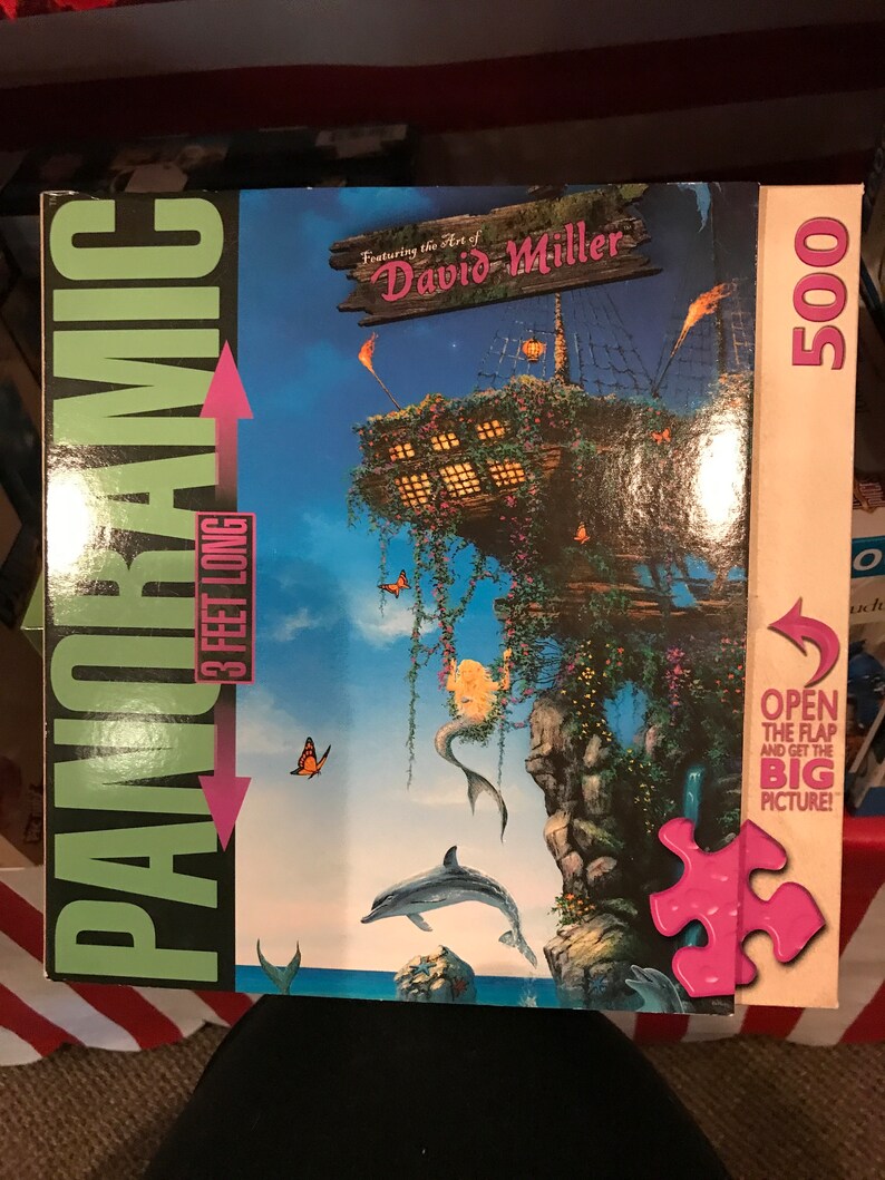 May include: A 500-piece panoramic jigsaw puzzle featuring the art of David Miller. The puzzle depicts a whimsical scene with a mermaid, a dolphin, butterflies, and a treehouse-like structure on a cliff overlooking the ocean. The box is green and black with the words "Panoramic" and "3 feet long" printed on it.