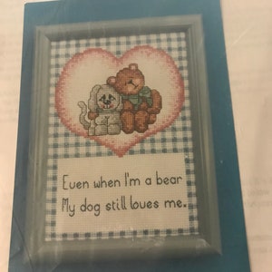 May include: Cross stitch pattern featuring a dog and a bear inside a heart. The text reads "DOG LOVES ME" and "Even when I'm a bear, My dog still loves me."