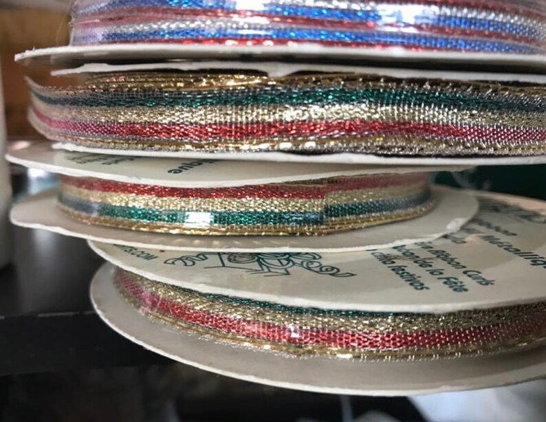 May include: Spools of metallic ribbon in various colors, including red, gold, green, and blue. The ribbon is stacked on white cardboard spools, with the edges of the ribbon visible. The ribbon appears to be for crafting or gift wrapping.