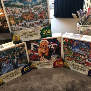 May include: Several Signature Collection 300-piece jigsaw puzzles. The puzzles feature winter scenes, including a snowy village, a deer in a forest, and puppies wearing Santa hats. The boxes are white with gold accents.
