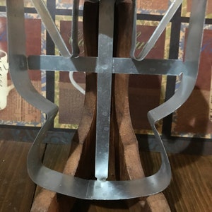 Puede incluir: Un cortador de galletas de metal plateado con forma de ángel con las alas extendidas y una cruz en el centro. El cortador se muestra en un soporte de madera.