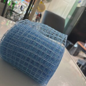 Puede incluir: Un rollo de cinta de malla decorativa de color azul claro con una textura de rejilla y sutiles hilos metálicos tejidos en el material. La malla es semitransparente y estructurada, mostrando un patrón cuadrado consistente en toda la superficie.