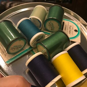 May include: A collection of spools of thread in various colors, including green, blue, and yellow, are arranged in a silver tray. The thread spools are labeled "Dual Duty" and "Coats & Clark Inc."