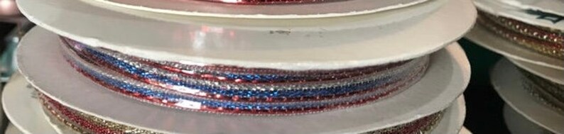 May include: Spools of decorative ribbon in red, white, and blue. The ribbon is spooled on white cardboard discs. The image shows a stack of spools, with the ribbon visible on the edges. The ribbon appears to be glittery.