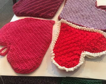 May include: Four crocheted heart-shaped pot holders in pink, red, purple, and white. The pot holders are on a white surface.