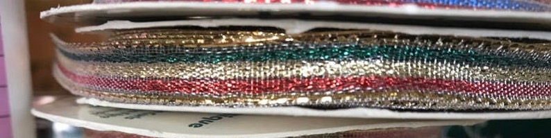 May include: Spools of decorative ribbon in various colors, including gold, red, green, and blue. The ribbon has a metallic sheen and a woven texture. The spools are stacked on top of each other, showing the different colors and textures.