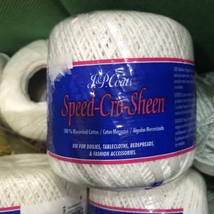 May include: A close-up of white J&P Coats Speed-Cro-Sheen thread spools. The label is blue with pink and white text, stating the thread is 100% mercerized cotton and suitable for doilies, tablecloths, bedspreads, and fashion accessories.