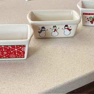 Puede incluir: Tres fuentes rectangulares de cerámica para hornear con interiores blancos y diseños festivos navideños. Una fuente presenta copos de nieve rojos, otra muñecos de nieve y la tercera bastones de caramelo y acebo.