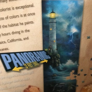 May include: A panoramic jigsaw puzzle featuring a lighthouse with a yellow light, a stormy sea, and the text "Having Fun One Piece at a Time".