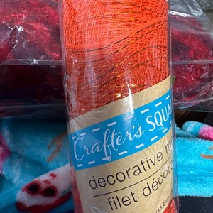 Puede incluir: Un rollo de malla decorativa de color naranja brillante con hilos dorados metálicos, envuelto en plástico transparente. La etiqueta de papel dice Crafter's Square decorative mesh, con dimensiones de 15,2 cm x 4,57 m impresas en el frente.