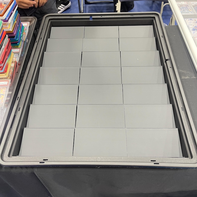 Pode incluir: Uma vitrine retangular cinza com v&aacute;rias prateleiras em camadas. A caixa est&aacute; aberta, revelando as prateleiras internas, projetadas para conter e exibir itens. A caixa est&aacute; em uma superf&iacute;cie preta.