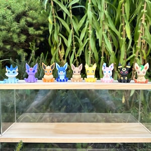 Può includere: Una collezione di nove figurine colorate, ognuna raffigurante un personaggio diverso con caratteristiche uniche. Le figurine sono disposte su una mensola di legno con un fronte in acrilico trasparente. Lo sfondo presenta un rigoglioso fogliame verde.