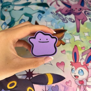 以下が含まれることがあります： 黒い縁取りとシンプルな笑顔の顔を持つ、紫色のメタモンの形をしたオブジェクト。背景には、グレイシア、エーフィ、ブラッキー、ニンフィアなど、さまざまなポケモンのキャラクターのカラフルなイラストが描かれており、ハートと星が描かれています。