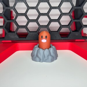 May include: A 3D-printed Diglett figure, a character from the Pok&eacute;mon franchise. The figure is brown with a pink mouth and black eyes, emerging from a gray rock formation. The background includes a black honeycomb-patterned structure and a red frame.